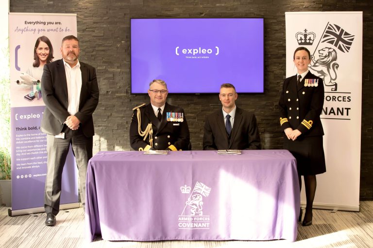 Four people, including two in military uniforms, stand and sit around a table with an "Armed Forces Covenant" cover. Behind them are banners and a screen displaying the "Expleo" logo in a bright office setting.