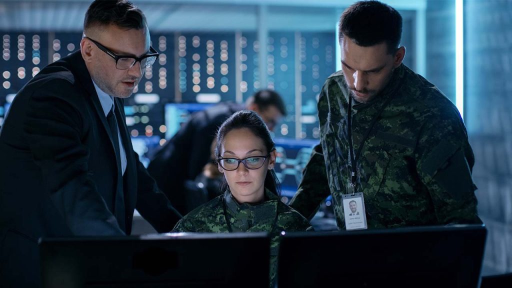 Three people in a dimly lit room focus on computer screens. Two are in military uniforms, and one is in a suit. They appear engaged in a discussion or analysis, with a backdrop of digital displays and equipment.