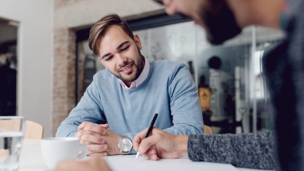 A man in a blue sweater with a watch smiles while sitting at a table, watching another person write on a piece of paper. The setting appears to be a casual indoor environment with soft lighting.