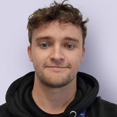 A person with short, curly brown hair and blue eyes is wearing a black hoodie. They are looking directly at the camera with a neutral expression. The background is a plain, light purple or gray color.