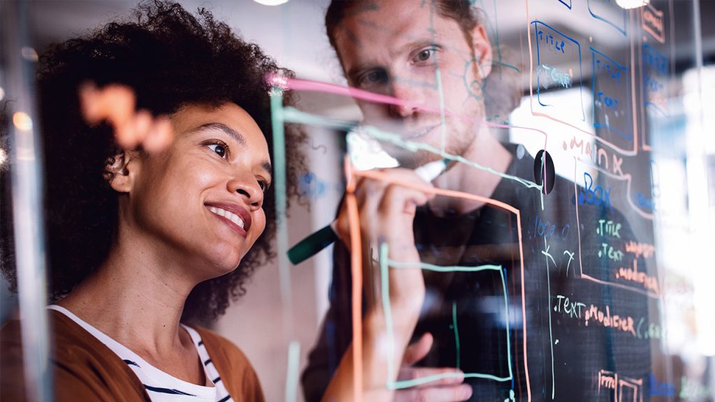 Two people collaborate, writing and drawing with colored markers on a transparent board covered with diagrams and notes. One smiles while observing the other's work. They appear engaged and focused in a creative environment.