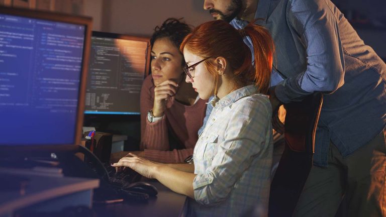 Three people work closely together at a desk with multiple computer monitors displaying code, focusing intently on the screens in a dimly lit room, suggesting a collaborative programming or IT environment.