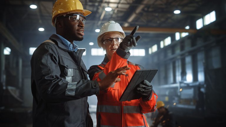 Two engineers in safety gear and helmets are standing in a factory. One is holding a tablet while both discuss and point to something out of view. The background shows industrial equipment and a spacious, well-lit environment.