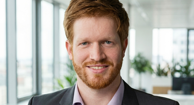 A man with short red hair and a beard, wearing a dark suit jacket and light purple shirt, smiles at the camera in a bright, modern office with large windows and plants in the background.