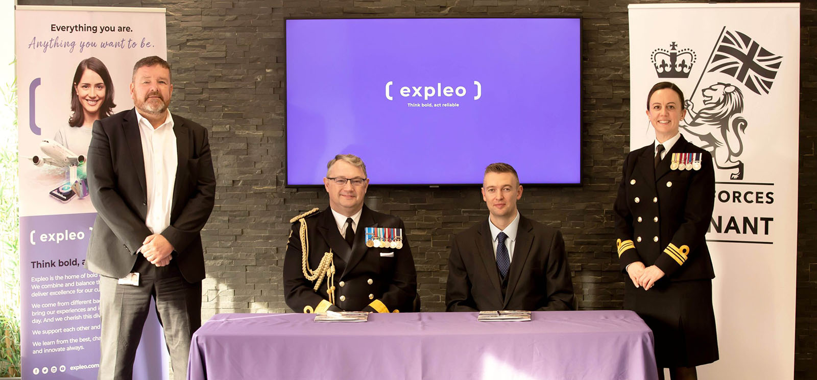 Four people pose at a table with a purple cloth. Two wear military uniforms with medals, two are in business attire. Behind them are banners and a screen displaying “expleo.” The setting appears formal, likely for a signing event.