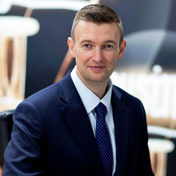 A man in a navy blue suit and polka dot tie sits and smiles at the camera, with a blurred office background behind him.