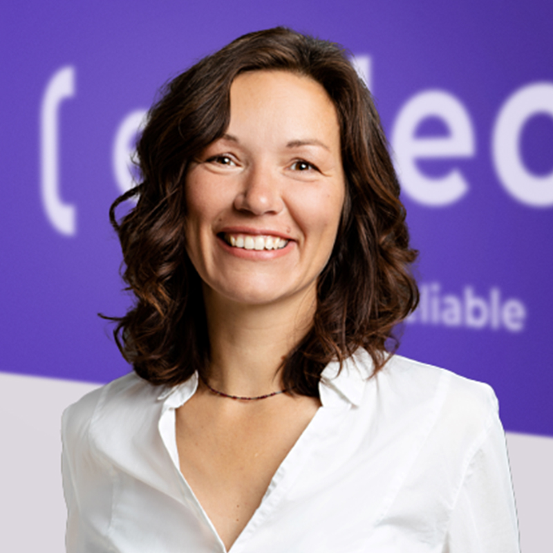 A woman with wavy brown hair, wearing a white blouse and a necklace, smiles in front of a purple background with partial white text.