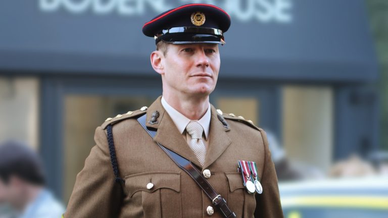 A man in a brown military uniform with medals and a peaked cap stands outdoors, looking forward with a serious expression. The background is blurred, showing part of a building and other people.