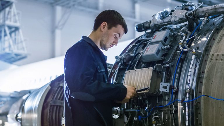 An aircraft maintenance technician in a navy blue jumpsuit, representing Expleo's commitment to excellence, inspects and works on the plane's engine inside a hangar. He is focused, using a tool on the intricate components, with the airplane and hangar structure as his backdrop.