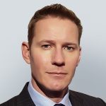 A man with short light brown hair wearing a dark pinstripe suit, light blue shirt, and tie, posing against a plain light gray background.