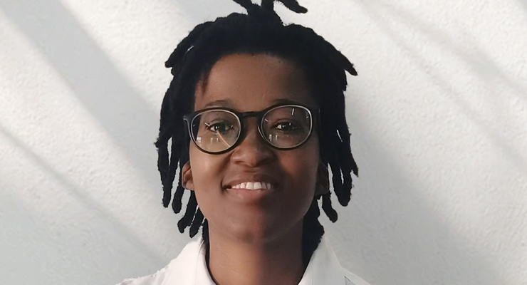 A person with short dreadlocks, wearing black glasses and a white shirt, smiles in front of a light-colored textured wall with shadow lines.