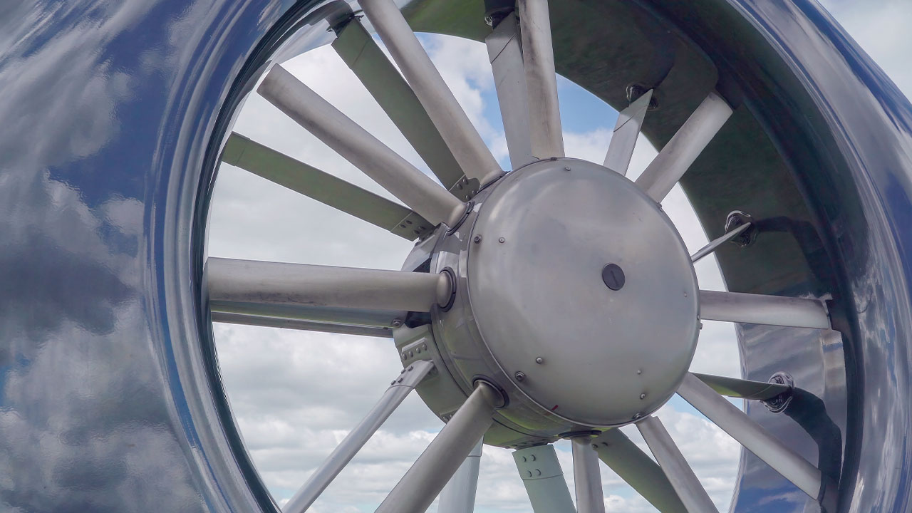 Close-up view of a large, metallic aircraft engine turbine with multiple blades, reflecting the cloudy sky.