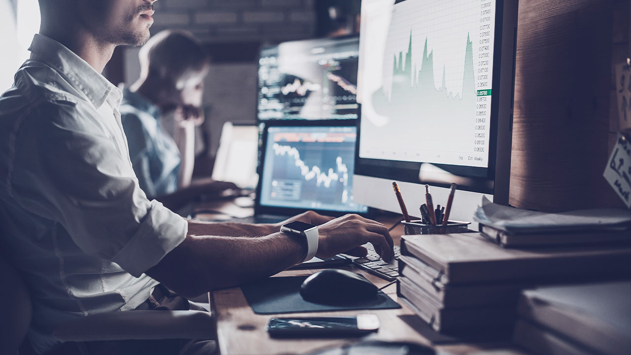 Two people work at desks with large monitors displaying stock charts and data. The workspace is cluttered with books, stationery, and a smartphone, creating a focused office environment.