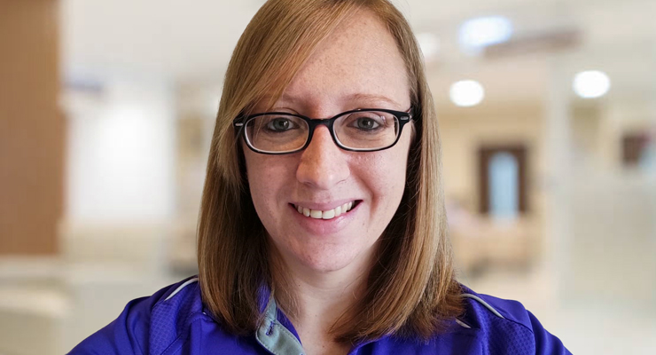 A woman with straight, light brown hair and glasses smiles at the camera. She is wearing a blue collared shirt and is in a softly blurred indoor setting.