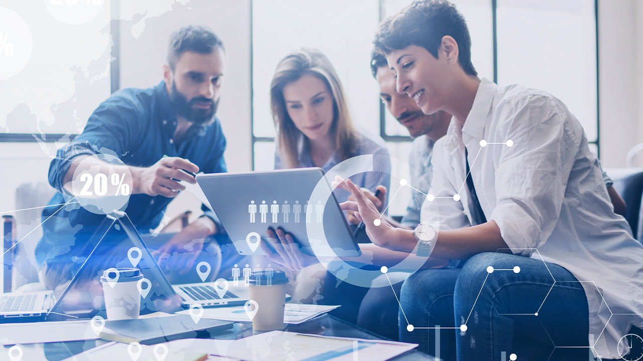 Four people sit together at a table, collaborating and looking at a laptop. Digital graphic overlays of data and network icons illustrate a technology and teamwork theme in a modern office.