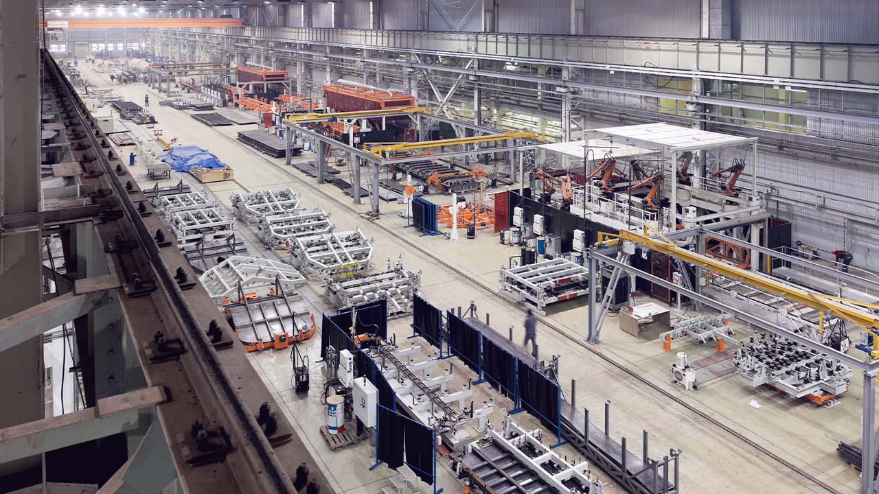 Large, brightly lit factory interior with assembly lines, industrial machinery, and workers scattered throughout. Overhead cranes and metal frameworks are visible, with various equipment and materials organized in sections.