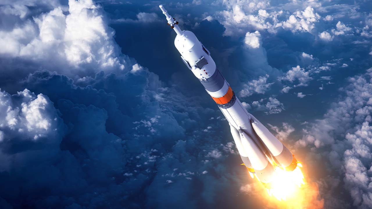 A rocket launches into the sky, trailing flames and smoke beneath it, while soaring above thick clouds in the atmosphere.