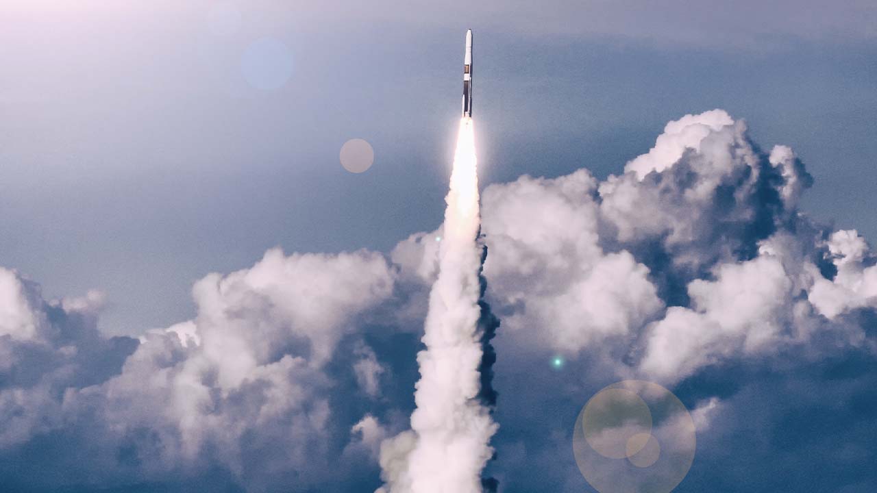A rocket launches into the sky, leaving a trail of smoke behind as it ascends through fluffy clouds under a blue sky with lens flares visible in the image.