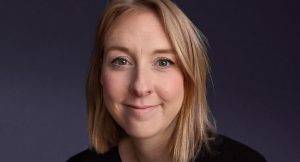 A person with shoulder-length blond hair smiles softly against a dark background. They are wearing a black top and have a light complexion.