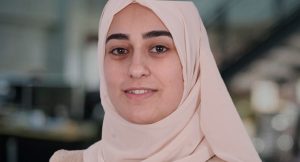A woman wearing a light beige hijab smiles softly while standing indoors. The background is blurred, showing a workspace with dark furniture and soft lighting.