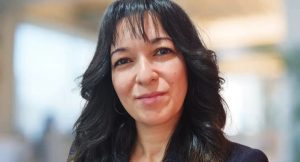 A person with shoulder-length dark hair and bangs smiles slightly at the camera. They are wearing a dark top. The background is blurred, suggesting an indoor setting with bright lighting.