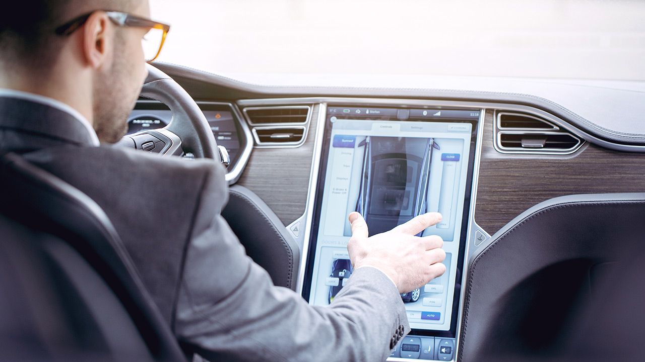 A person in a suit sits in a car, interacting with a large touchscreen display on the center console, adjusting vehicle settings or navigation options.