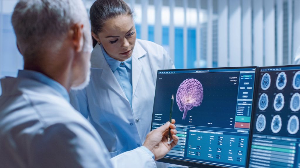 Two doctors in lab coats examine brain scans on a computer monitor. One doctor points at a 3D brain image on the screen while the other watches attentively. Additional brain scan images are visible on an adjacent monitor.