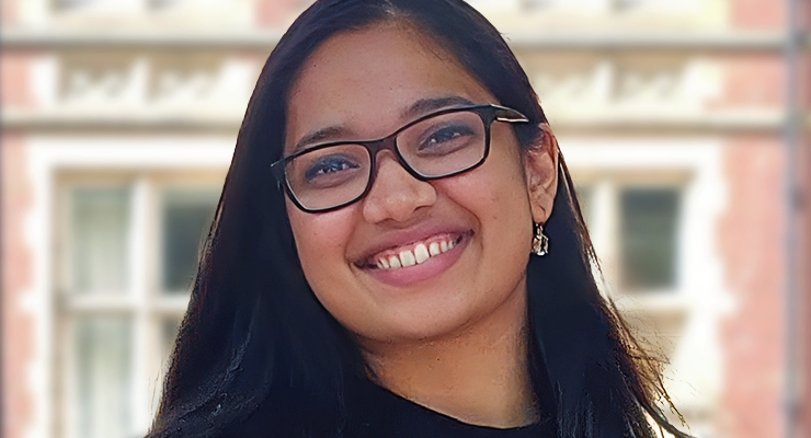 Smiling person with long dark hair and glasses, standing outdoors in front of a blurred building.