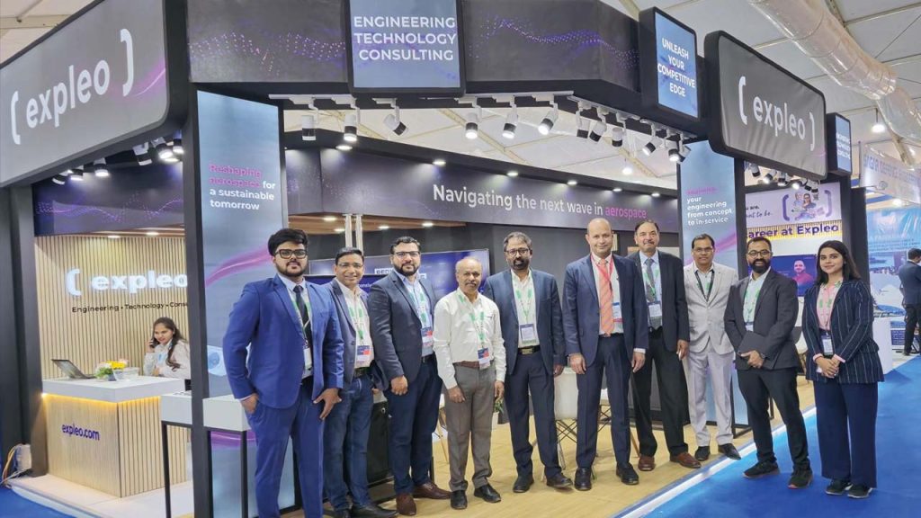 A group of ten professionally dressed people pose and smile in front of an Expleo booth at a technology expo, with signs reading "ENGINEERING TECHNOLOGY CONSULTING" visible above them.