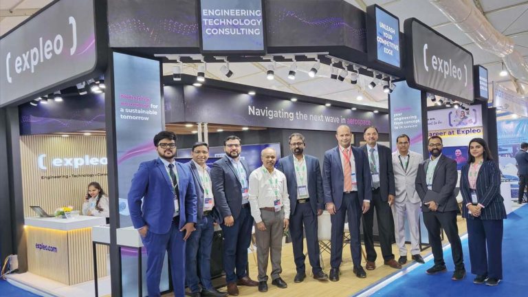 A group of ten professionally dressed people pose and smile in front of an Expleo booth at a technology expo, with signs reading "ENGINEERING TECHNOLOGY CONSULTING" visible above them.