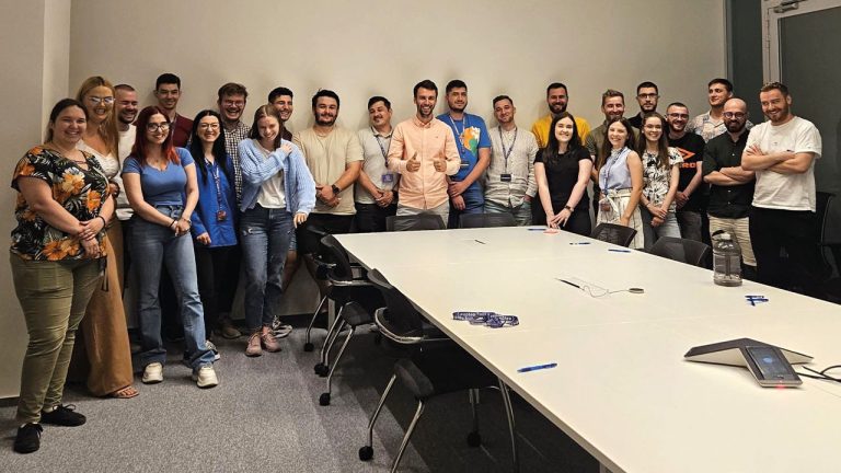 A large group of people stand smiling in a conference room around a long table. They are casually dressed and appear to be in a meeting or workshop setting. The room has plain walls and several chairs are placed around the table.