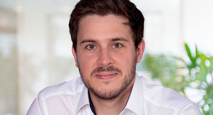 A man with short brown hair and a trimmed beard, wearing a white collared shirt, sits indoors with a blurred background and green plants visible behind him.