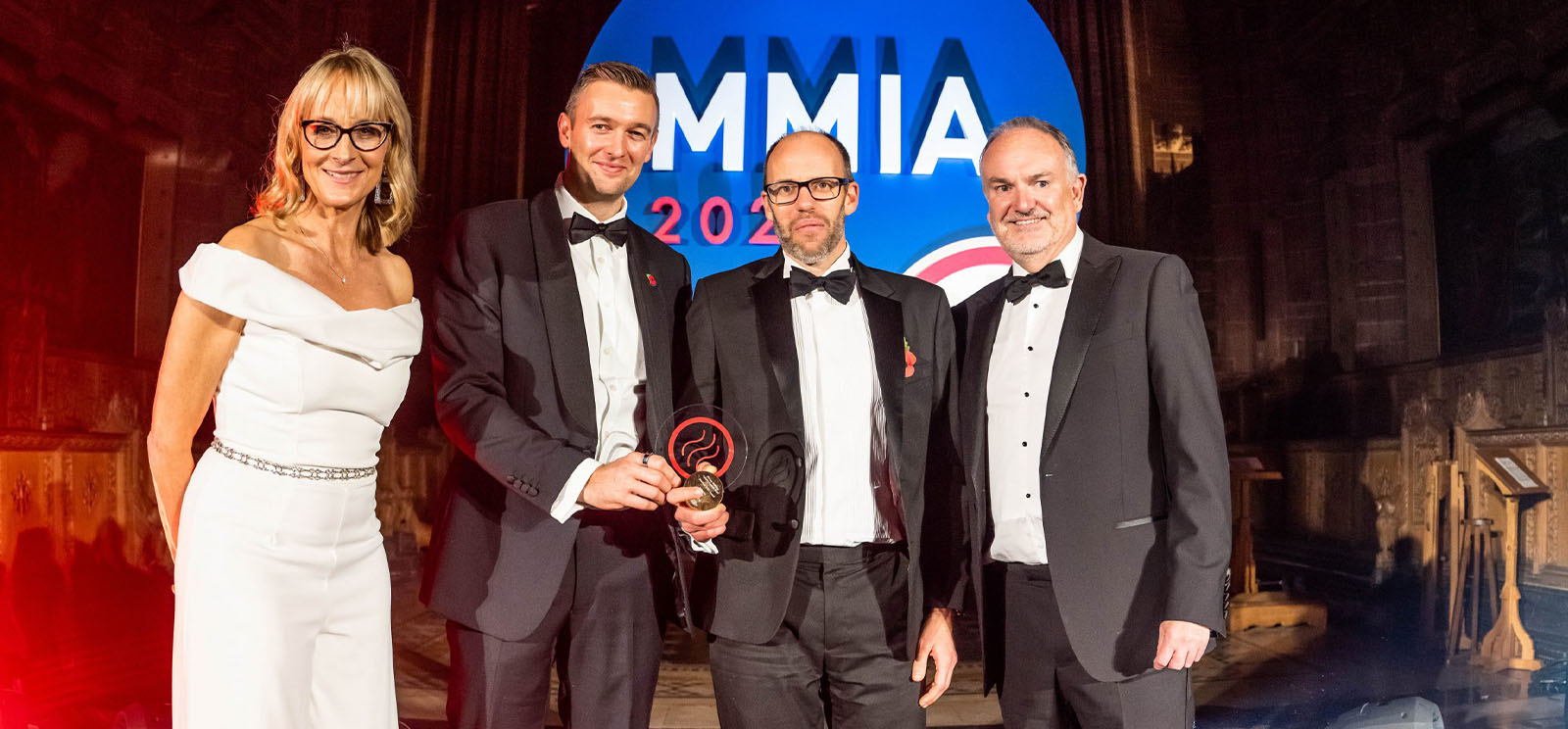 Four people in formal attire stand smiling at an awards event, with two men in the center holding a trophy. A large blue MMIA 2021 sign is illuminated in the background.