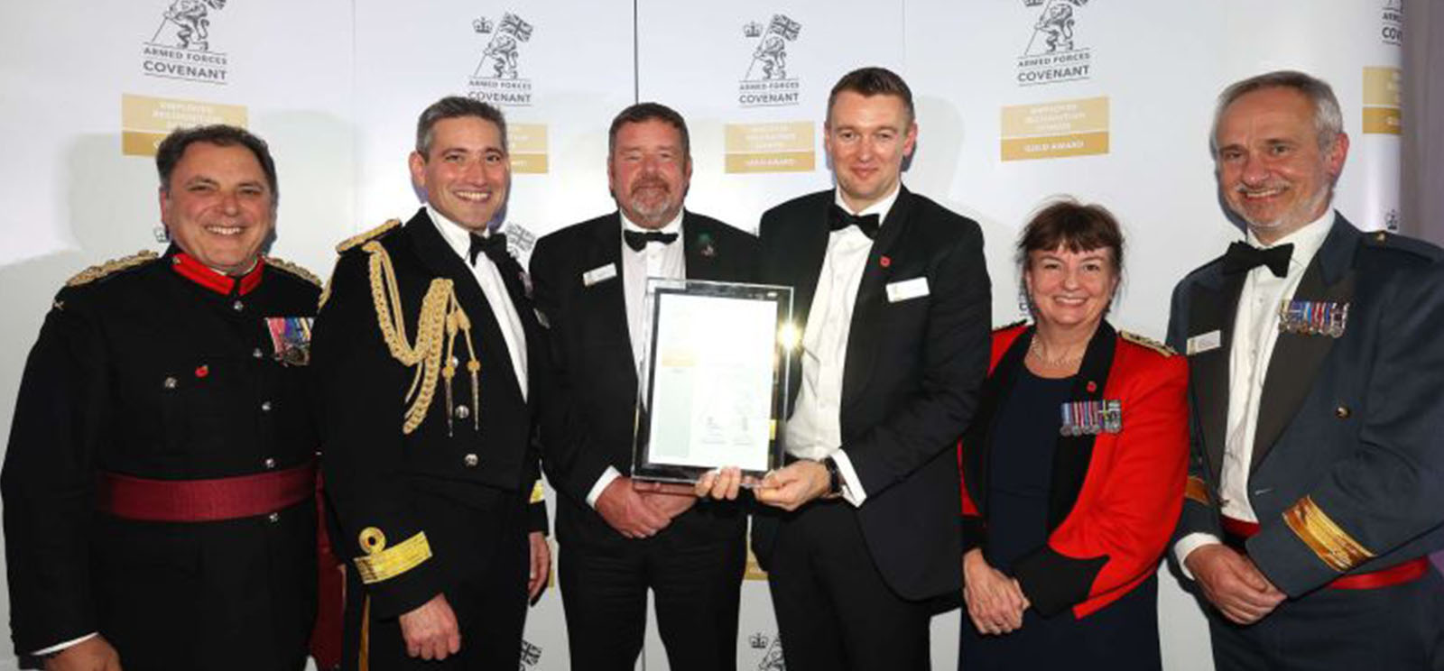 Six people in formal military attire and evening wear stand together, smiling, as two of them hold a framed award certificate. A backdrop behind them displays the Armed Forces Covenant logo.