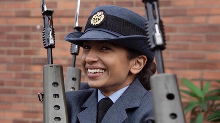 A smiling woman in a blue military uniform and hat stands between two rifles held upright, with a brick wall in the background.