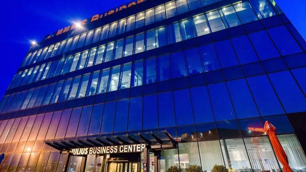 Modern glass office building at dusk, with interior lights glowing. The entrance sign reads "Julius Business Center." Reflections and blue sky create a sleek, professional atmosphere.