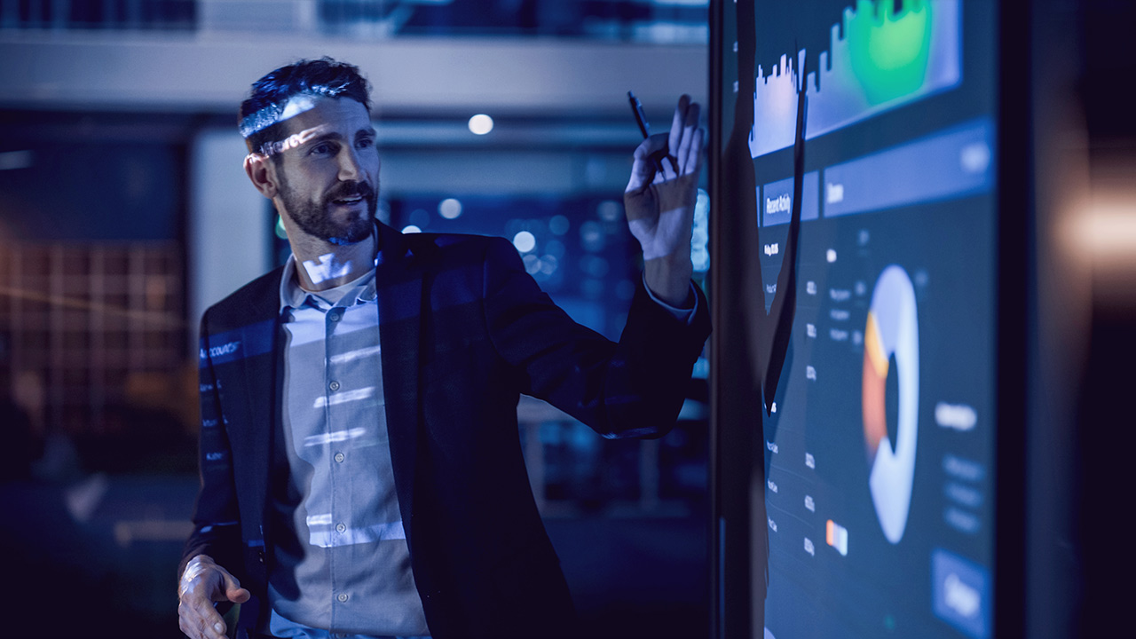 A man in a suit stands in a dark room, pointing at a large digital screen displaying colorful data charts and graphs, illuminated by the projected visuals.