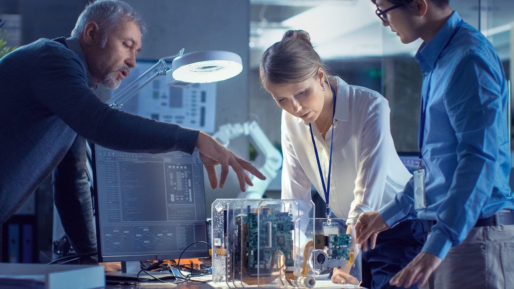 Three engineers collaborate around a desk, examining electronic components and circuit boards—reflecting the innovation found in our energy and utilities roles—in a modern office or lab with technical diagrams on a computer monitor.