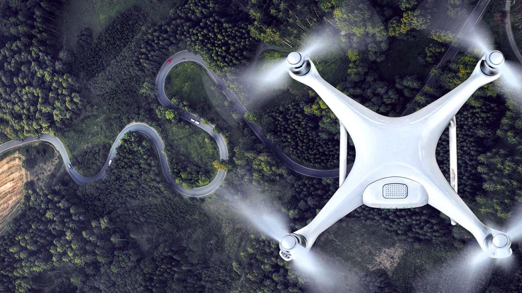 A white drone flies above a winding road surrounded by dense green forest, capturing an aerial view of the curvy path and landscape below—reflecting the innovative perspective found in our energy and utilities roles.