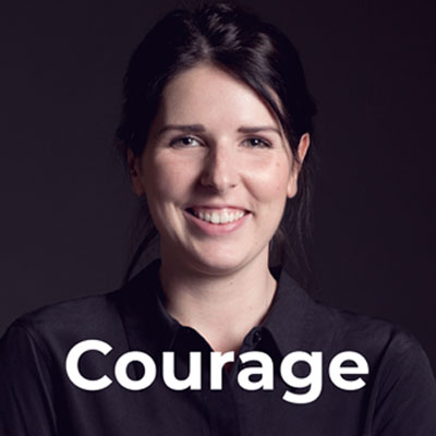 A smiling woman with dark hair in a black shirt stands against a dark background. The word "Courage" is written in bold white letters across the lower part of the image.