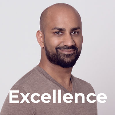 A smiling man with a beard and shaved head wearing a beige sweater stands against a light background. The word "Excellence" is written in large white letters at the bottom of the image.