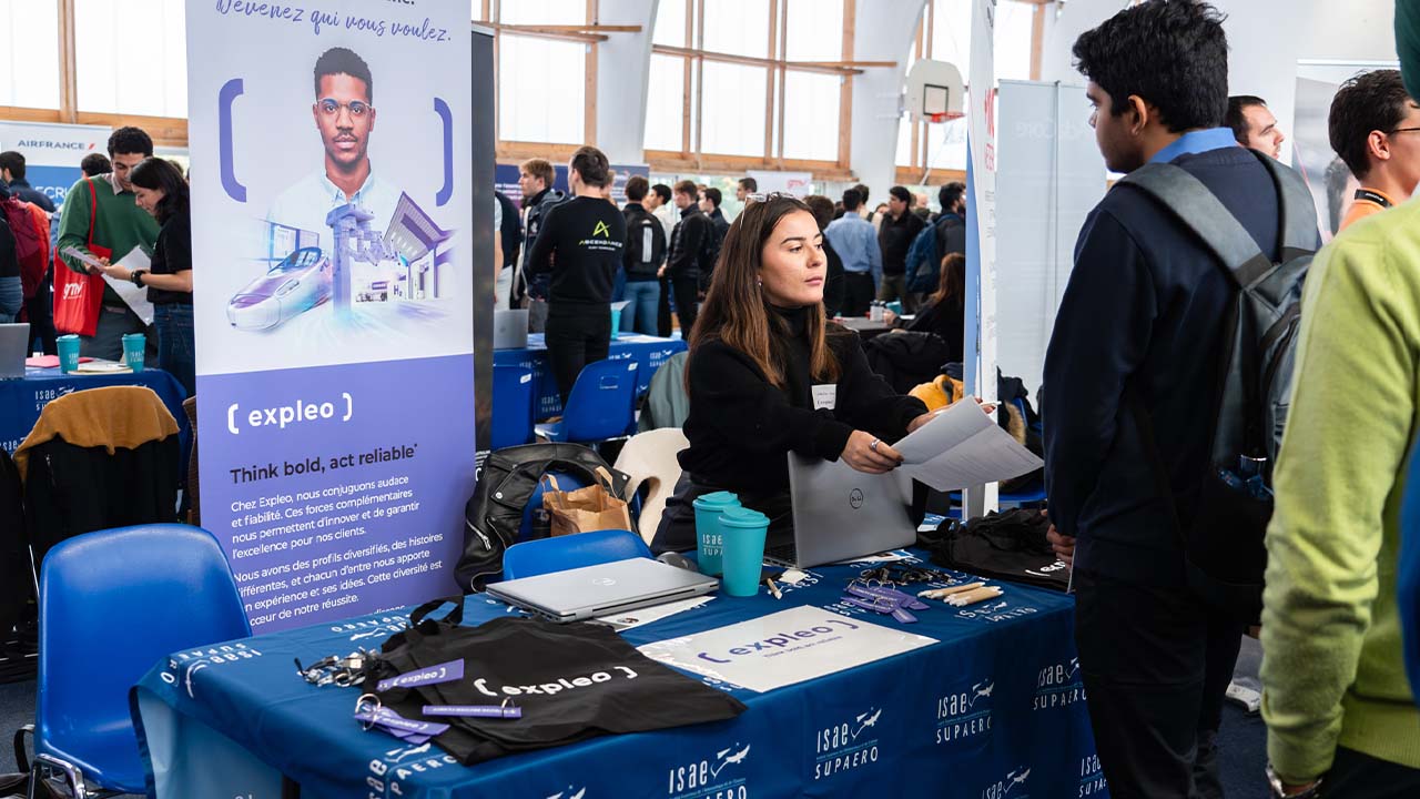 A woman at a career fair booth talks to a man. The booth, branded with “Expleo,” has brochures, tote bags, and pens on the table. Other job seekers and company displays are visible in the background.