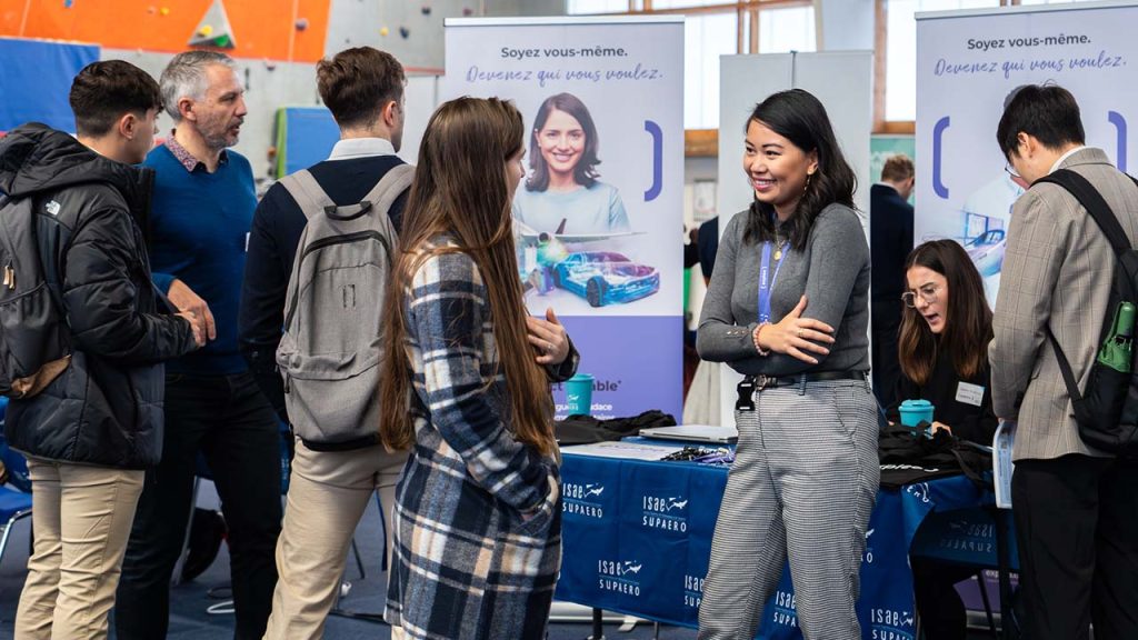 A group of people talk and network at a career fair booth. Two women are smiling and conversing in front of promotional banners and tables with informational materials. Other attendees interact nearby.