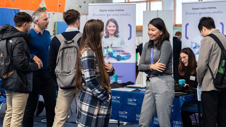 A group of people talk and network at a career fair booth. Two women are smiling and conversing in front of promotional banners and tables with informational materials. Other attendees interact nearby.