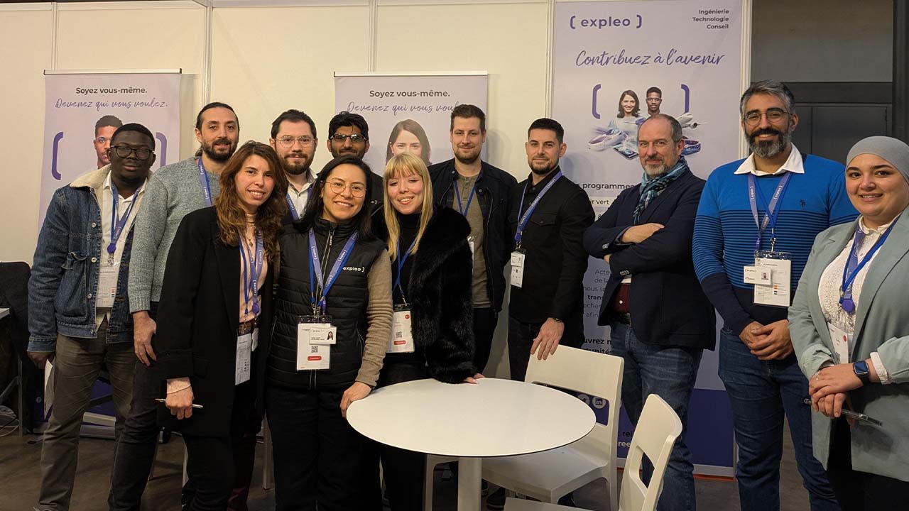 A group of people wearing business badges stand and smile for a photo at an event. Behind them are informational banners and a white table with chairs in the foreground.