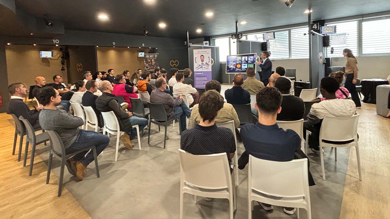 A group of people sit on white chairs facing a presenter speaking at the front of a modern room, with presentation slides displayed on screens and banners set up behind the speaker.