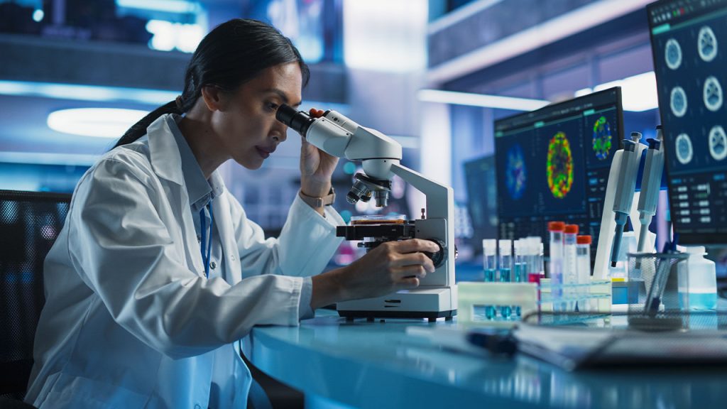 A scientist in a lab coat looks through a microscope at a laboratory workstation, surrounded by test tubes and computer monitors displaying colorful scientific images.