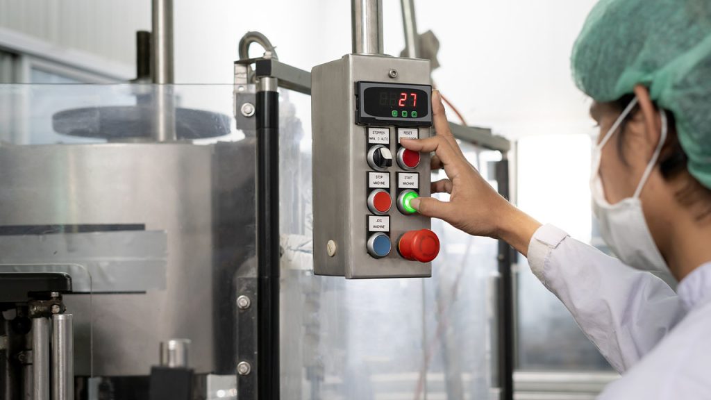 A person wearing a lab coat, hairnet, and face mask operates an industrial machine by pressing buttons on a control panel with a digital display showing the number 27.