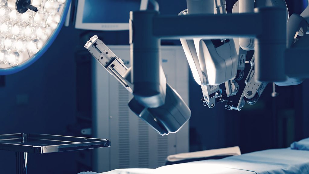 A close-up view of robotic surgical arms positioned over an operating table in an operating room, with medical equipment and monitors visible in the background.