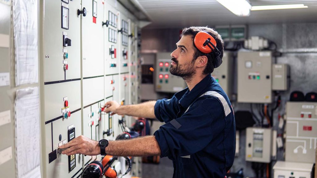 A man in a navy blue uniform and orange ear protection is examining a control panel in an industrial setting. He is surrounded by various switches and buttons, and appears focused on adjusting a component.